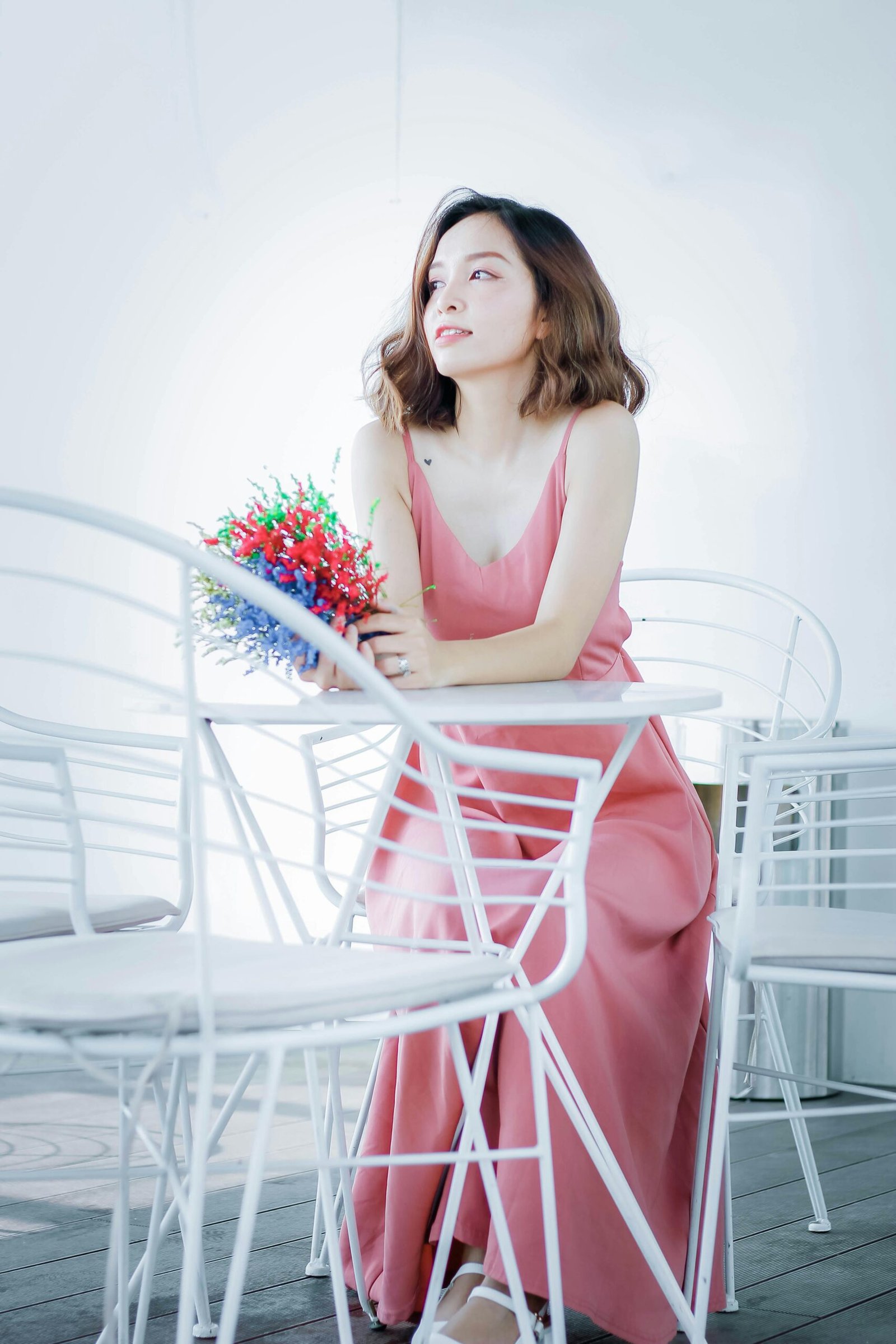 A woman in a pink dress holding colorful flowers while sitting indoors on white chairs.