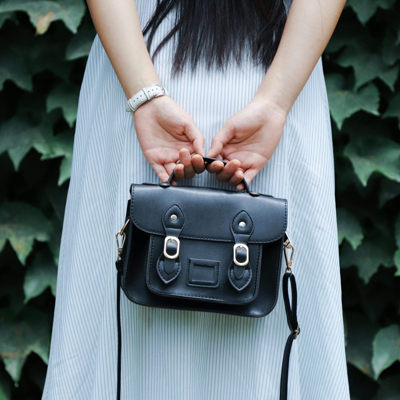 A woman in a striped dress holds a black leather handbag with lush green leaves in the background.